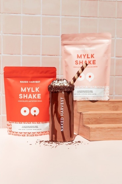A clean, studio-lit shot of two product bags flanking a chocolate lactation shake, utilizing a consistent soft-pink and vibrant orange color palette.